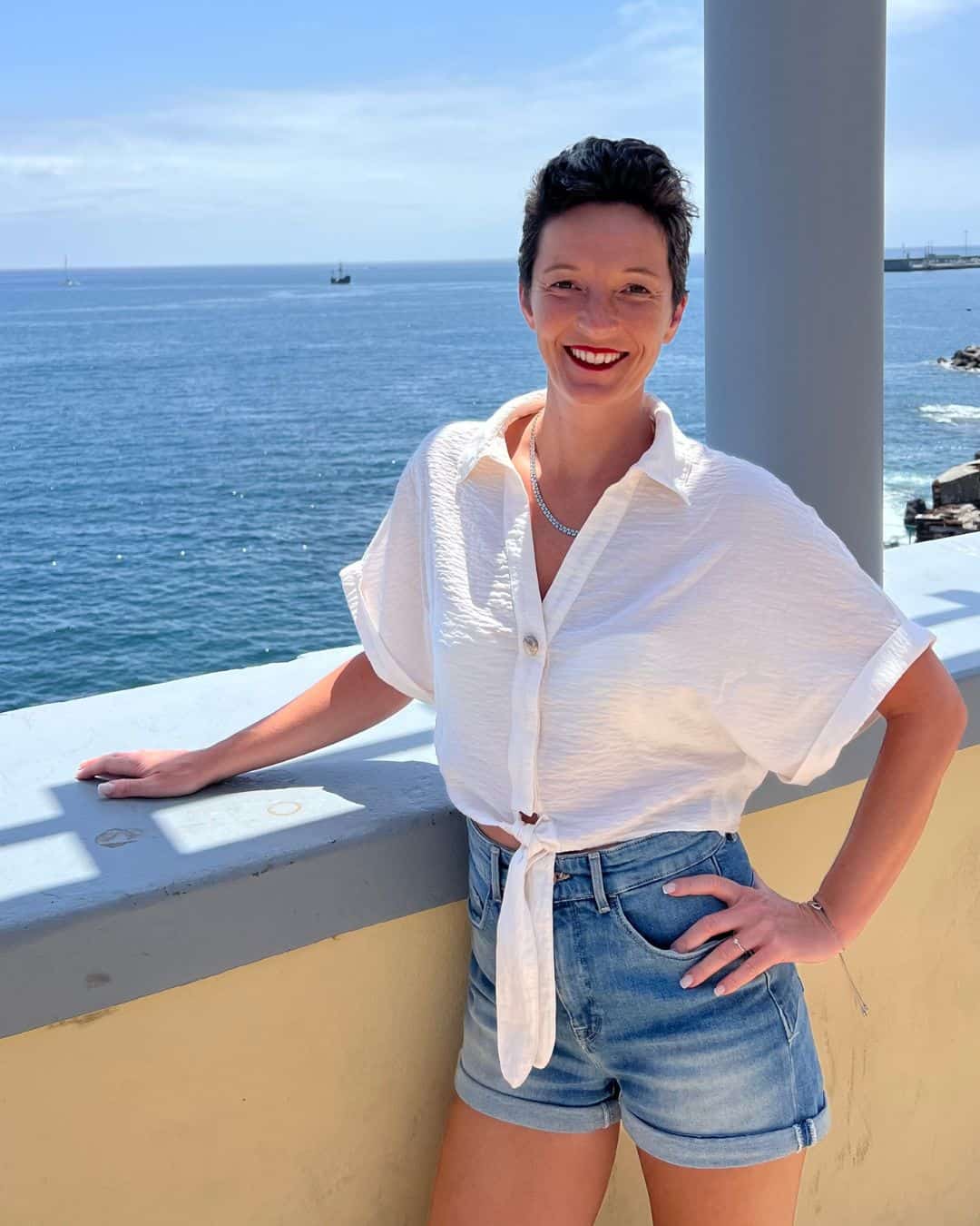 Frau steht lächelnd auf einer Terrasse mit Meerblick – Technik-Begleiterin für Onlinekurse und Onlinebusiness im DACH-Raum.