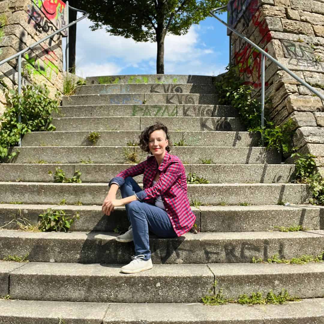 Frau sitzt entspannt auf einer Steintreppe im Freien und lächelt in die Kamera – Technik-Begleiterin für Onlinekurse und Onlinebusiness.