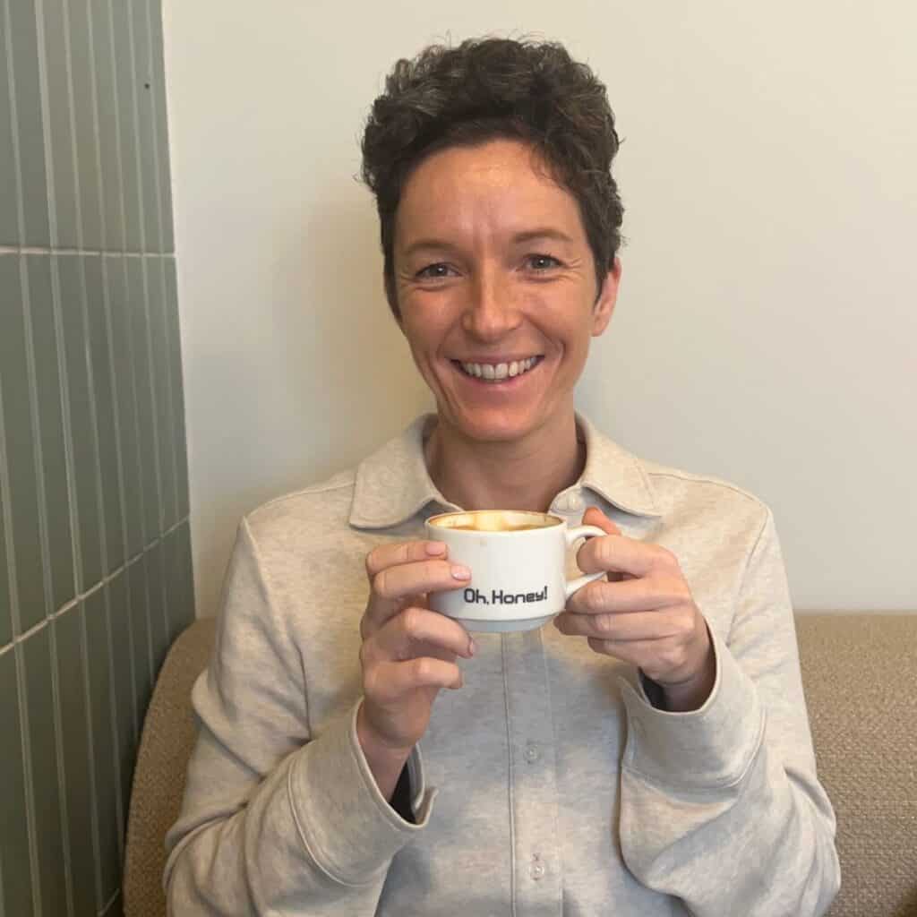 Frau sitzt entspannt in einem Café und hält lächelnd eine Tasse Kaffee in den Händen – Technik-Begleiterin für Onlinekurse und Onlinebusiness.